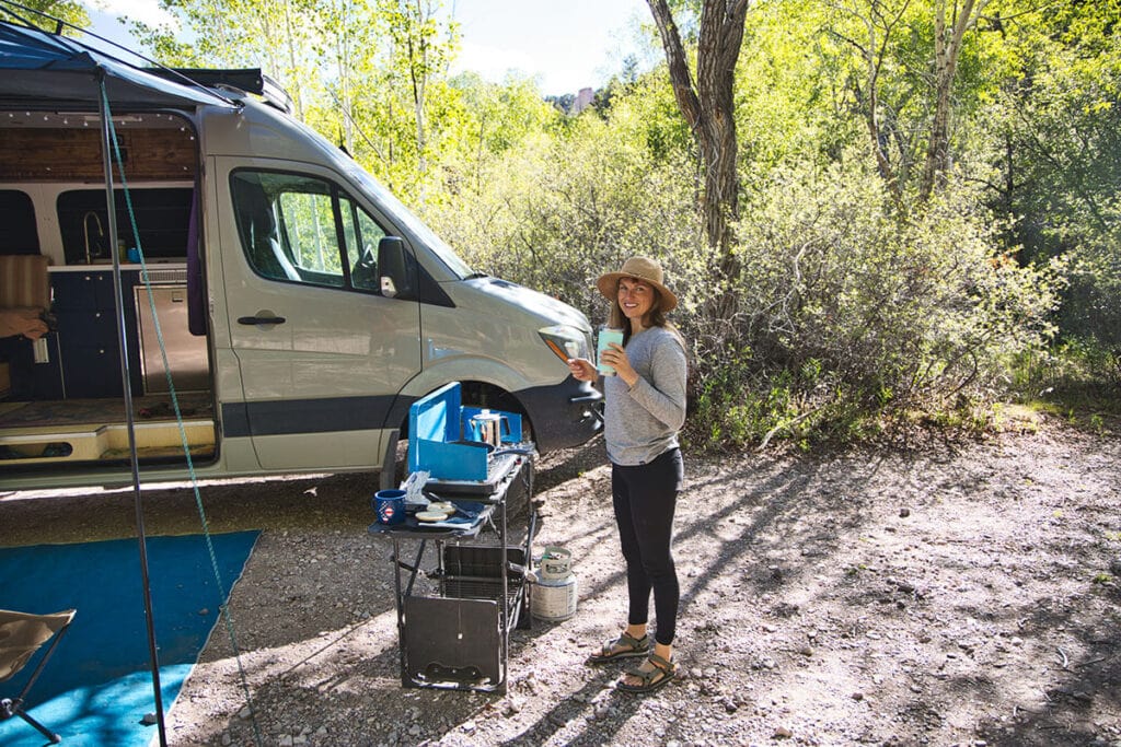 A woman standing outside of her Sprinter van cooking on a propane camp stove